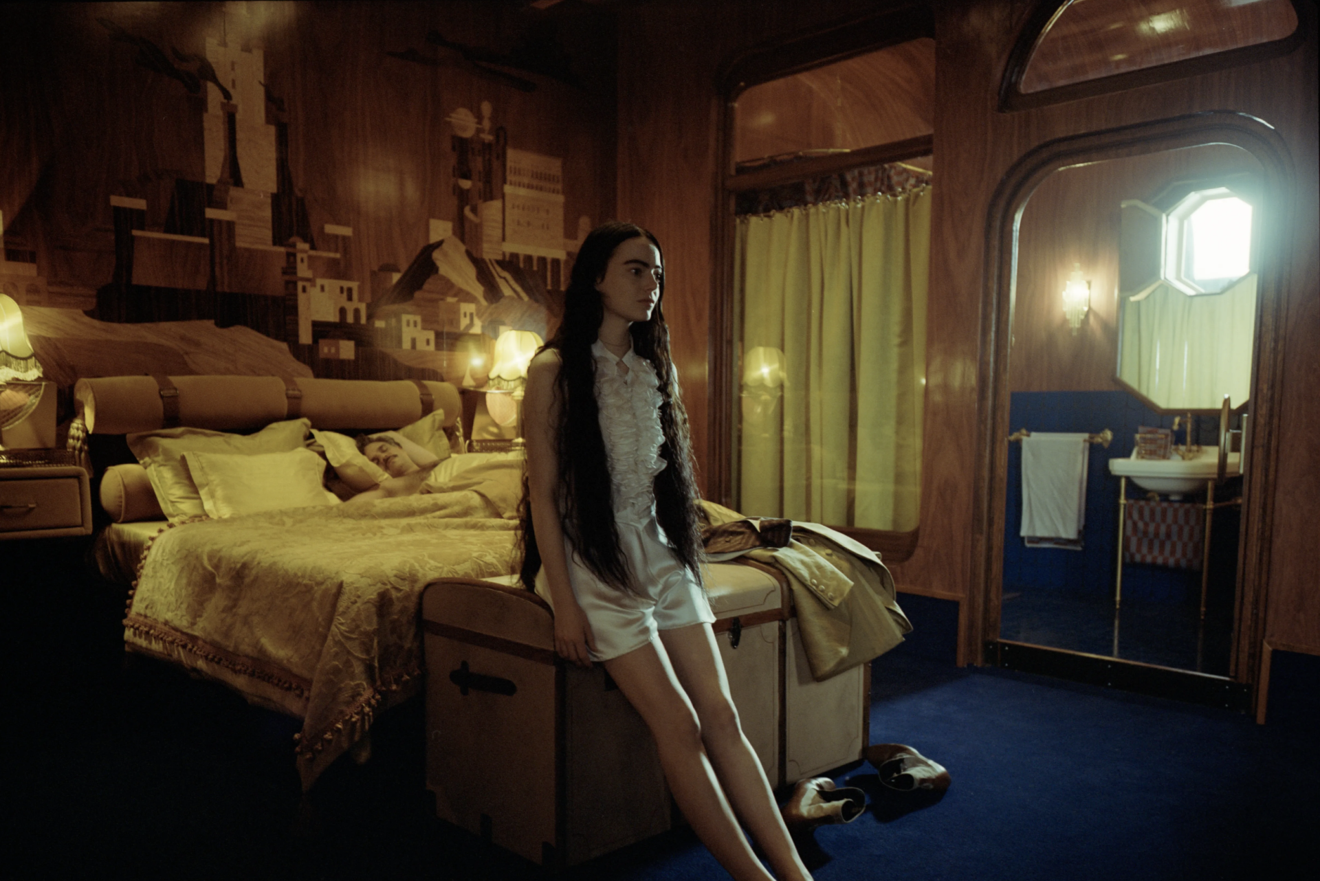 Imagem do filme 'Pobres Criaturas', onde Bella, uma garota de cabelos longos e pretos, está olhando fixamente para o horizonte, sentada em um baú na frente de uma cama em um quarto de luz amarela. 