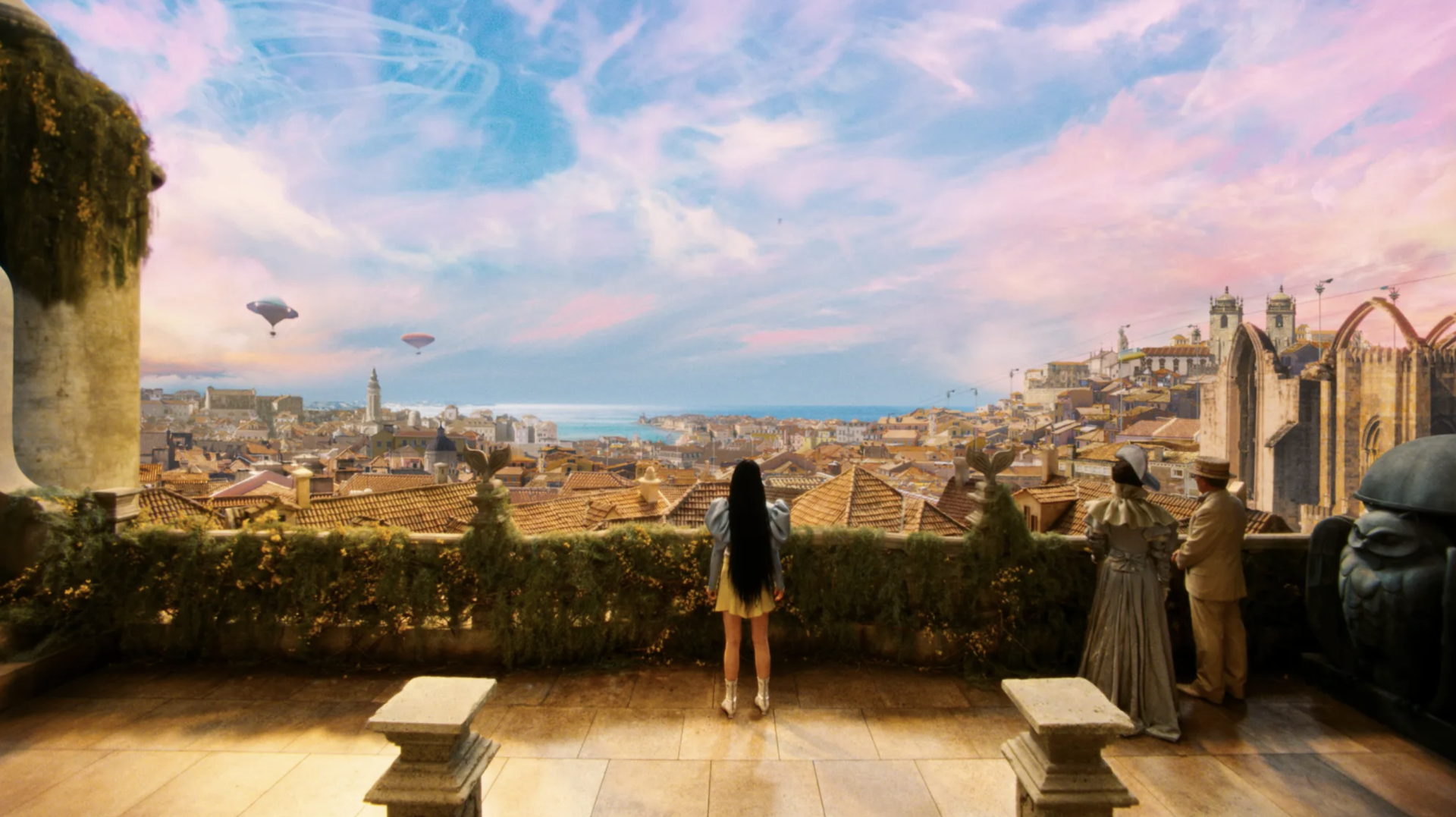 Imagem do filme 'Pobres Criaturas', onde Bella, com longos cabelos pretos, vislumbra uma Lisboa retrofuturista do alto de um miradouro, com céu azul e nuvens em tons rosa. 