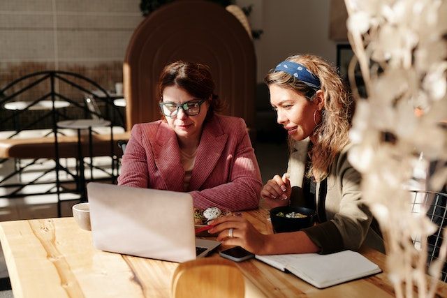 Two ladies setting around a table discussing the food act 2014 requirements displaed on a laptop scree. One lady is smilling she is  the food business operator who just opened her food business receivening an audit from the other lady who is the Enviromental Health Officer or council verifier. 