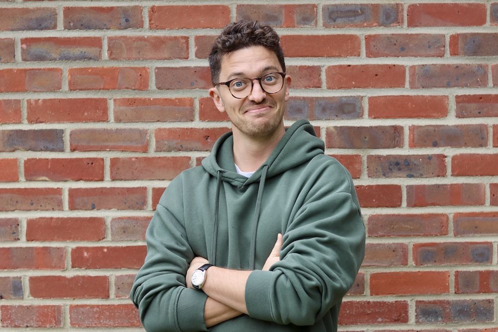Charlie with his arms crossed, wearing a green hoodie in front of a brick wall