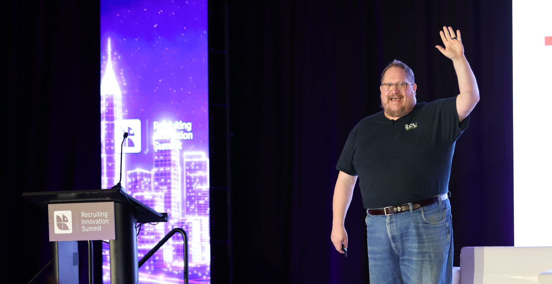 Shally Steckerl on stage at the ERE Recruiting Innovation Summit in Atlanta, smiling with one hand raised in a wave. He wears a dark shirt and jeans, with the summit podium beside him and a purple Atlanta skyline on the screen behind.