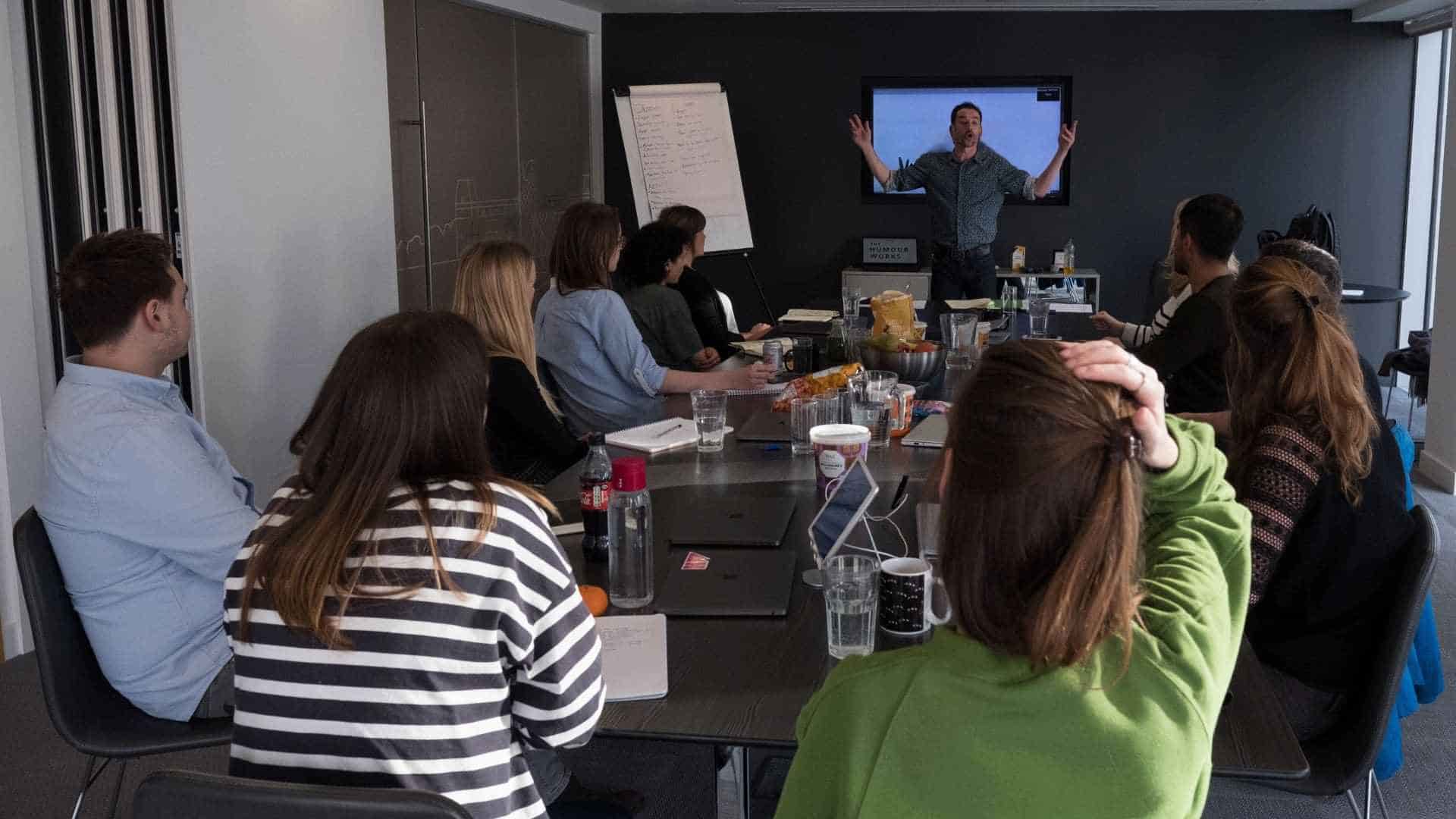Wide shot of Alfie at the far end of a workshop room with a dozen students in front of him as he addresses them with his arms raised wide